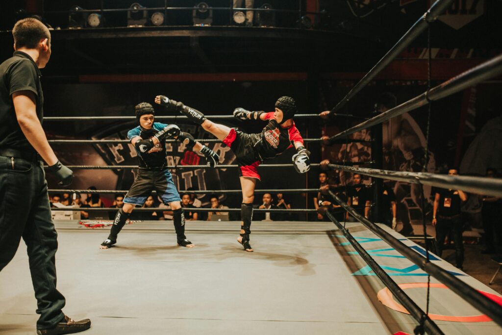 Boxing:
Boxer kicks opponent, who dodges and defends, furthermore referee watches closely, commentators and judges also observe from ringside.