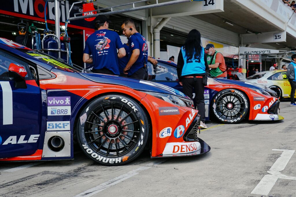 Sports Cars lined up in garages, preparing for racing event, teams and crew members surrounding vehicles.