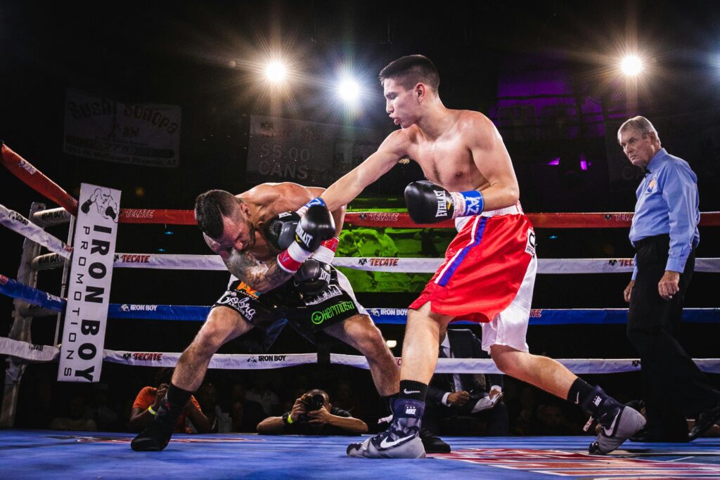 Boxing:
Boxer lands powerful punch on opponent's chest, sending them bending down, referee also watches in surprise, under bright ring lights.